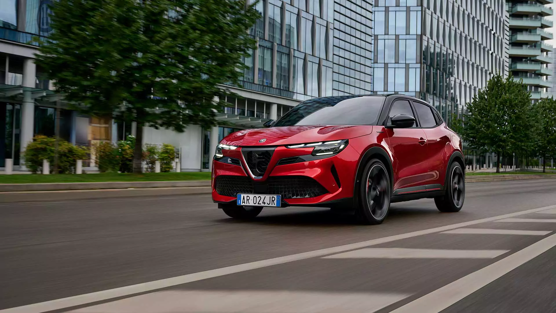 Ein roter Alfa Romeo Geländewagen fährt auf einer von modernen Glasgebäuden und Bäumen gesäumten Stadtstraße und überquert bei Tageslicht die Zebrastreifen.