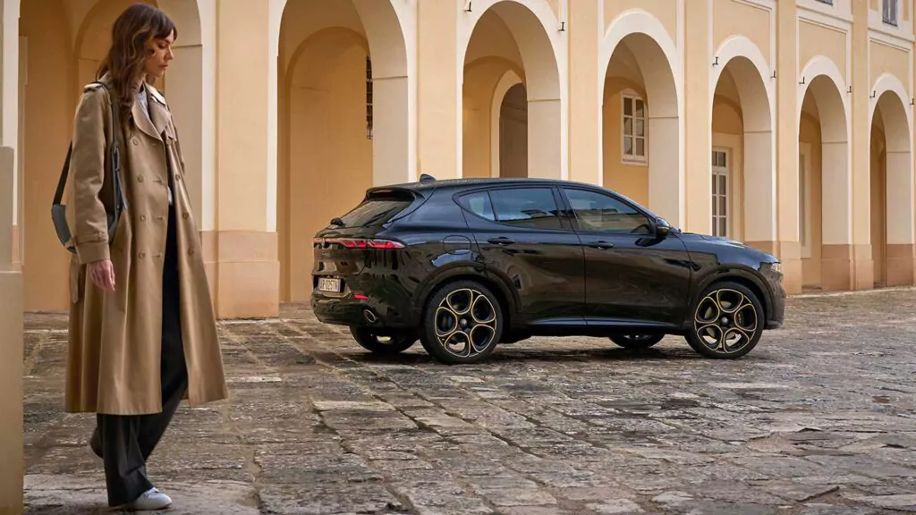 Eine Frau in einem hellbraunen Trenchcoat geht an einem schnittigen schwarzen Alfa Romeo Tonale Intensa vorbei, der auf einem kopfsteingepflasterten Innenhof vor einem hellen historischen Gebäude mit großen Bögen geparkt ist.