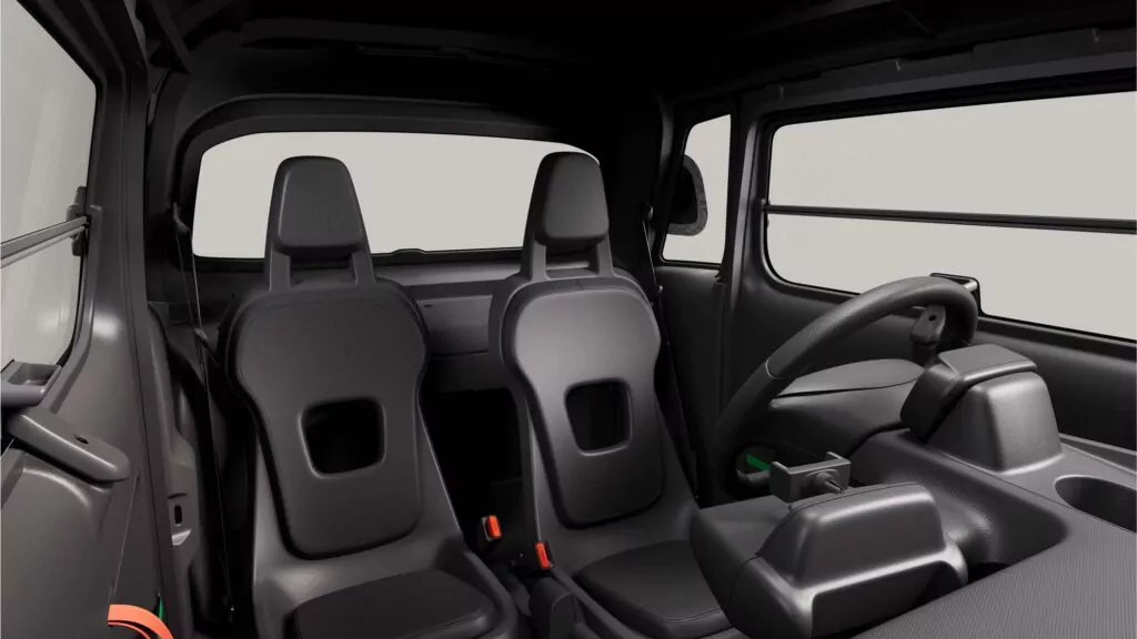 Das Bild zeigt den Innenraum eines Kleinwagens mit zwei minimalistischen schwarzen Sitzen, einem einfachen Lenkrad und einem spärlichen Armaturenbrett, alles in einem modernen, utilitaristischen Design. Der Hintergrund ist hellgrau und hebt das Innere des Fahrzeugs hervor.