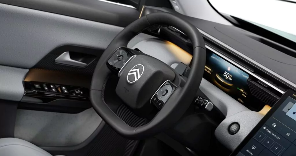 Nahaufnahme des Innenraums eines modernen Fahrzeugs mit dem Lenkrad mit Citroën-Logo, dem digitalen Armaturenbrett mit Geschwindigkeitsanzeige, einem Teil des Infotainment-Bildschirms und den Türsteuerungen.