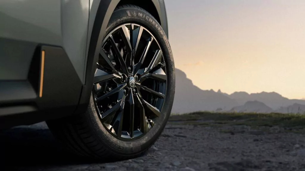 Nahaufnahme eines glänzenden schwarzen Autorads und Reifens auf einem felsigen Boden mit Bergen und einem weichen, orangefarbenen Himmel im Hintergrund bei Sonnenuntergang.