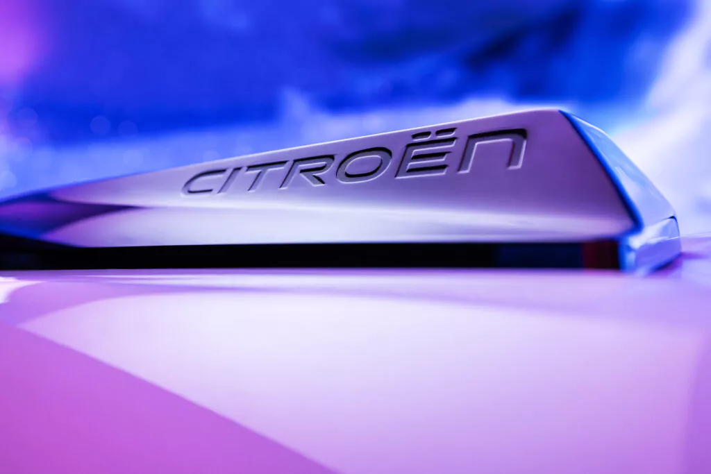 Nahaufnahme eines metallischen Citroën-Emblems auf einer glatten, geschwungenen Oberfläche des Citroën e-C3 Aicross, mit einem verschwommenen blauen und violetten Hintergrund, der den Markennamen in erhabenen Buchstaben hervorhebt.