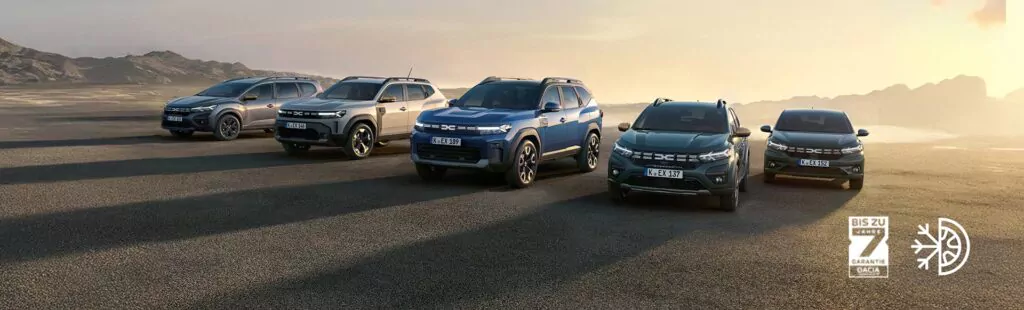 Fünf Dacia Geländewagen parken nebeneinander auf einer offenen, gepflasterten Fläche bei Sonnenuntergang mit Bergen im Hintergrund. Der Himmel ist klar, und zwei Dacia-Logos sind auf der rechten Seite des Bildes zu sehen.