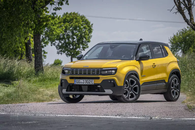 Ein leuchtend gelber Jeep-Geländewagen mit schwarzen Akzenten parkt auf einer Schotterstraße, umgeben von Gras und Bäumen, unter einem klaren blauen Himmel.