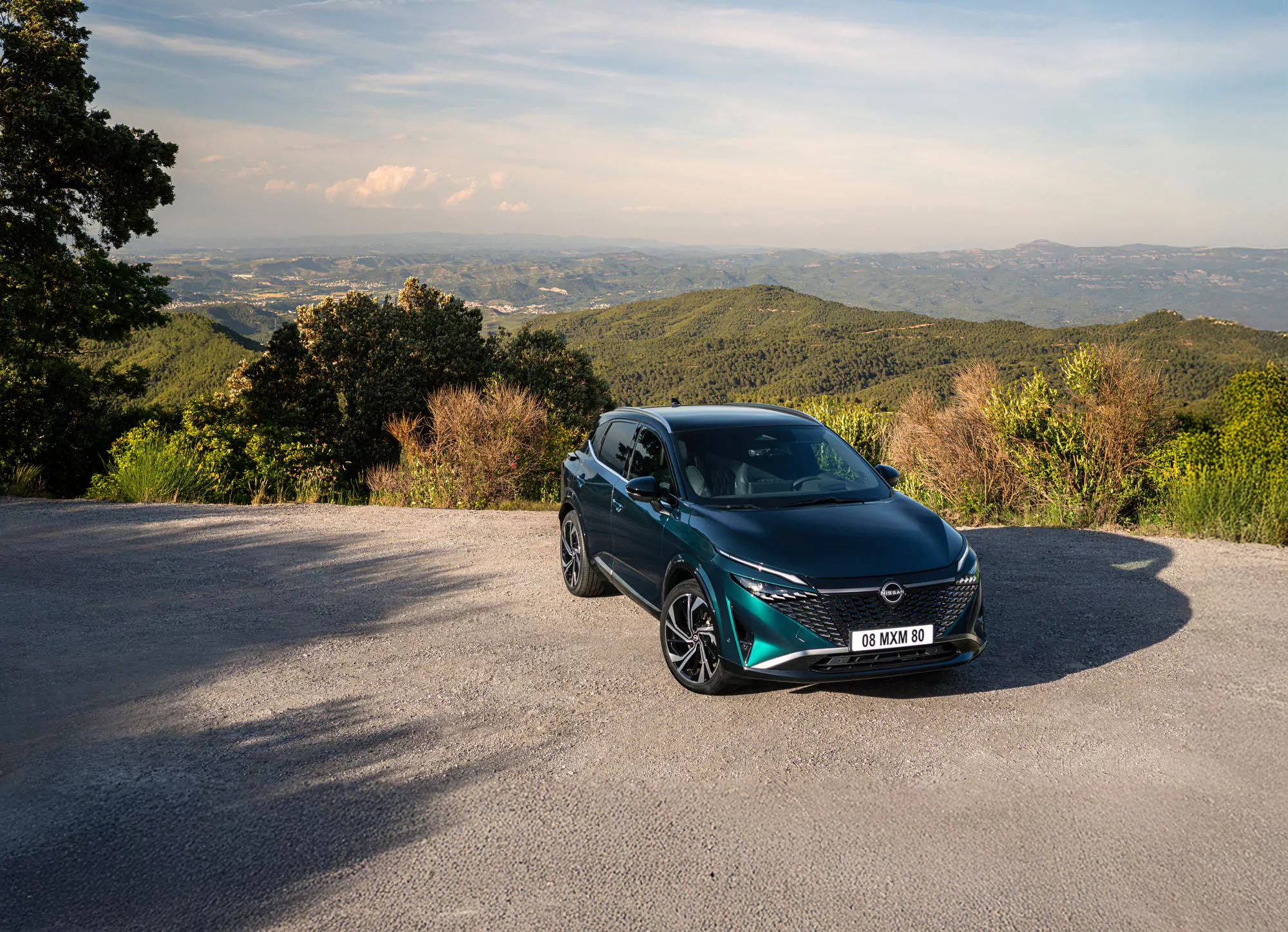 Ein blaugrüner Peugeot 308 ist auf einer Schotterfläche mit grünen Hügeln und einem wolkenverhangenen Himmel geparkt. Bäume und Büsche umgeben das Auto und schaffen eine malerische Landschaft, die sich perfekt für eine Erkundungstour mit dem Nissan Qashqai TEKNA eignet.