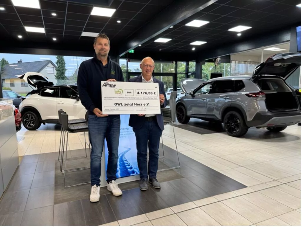 Zwei Männer stehen in einem Autohaus und halten einen großen Scheck über 1.476,93 Euro in der Hand, ausgestellt auf OWL zeigt Herz e.V. Hinter ihnen stehen zwei Autos mit offenen Hintertüren, große Fenster geben den Blick auf Häuser im Freien frei.