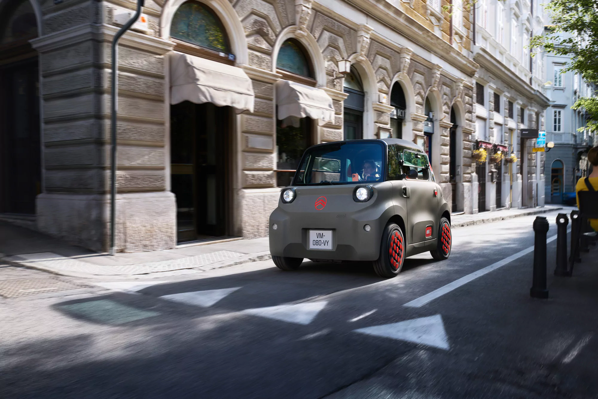 Ein kleines, modernes zweisitziges Elektroauto mit grauer Karosserie und roten Akzenten fährt durch eine schmale europäische Stadtstraße, die von historischen Gebäuden und Straßencafés gesäumt ist.