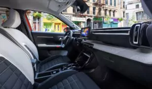 Ansicht des Innenraums eines modernen Autos von der Beifahrerseite aus, mit schwarzen und grauen Sitzen, einem digitalen Armaturenbrett, einem zentralen Bildschirm und einer Stadtstraße mit bunten Gebäuden, die durch die Fenster sichtbar sind.