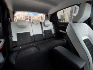 Das Bild zeigt das Innere eines modernen Autos, wobei der Schwerpunkt auf dem Rücksitzbereich mit schwarzen und hellgrauen Sitzen, Kopfstützen und einem Blick auf Gebäude durch die Heck- und Seitenfenster liegt.