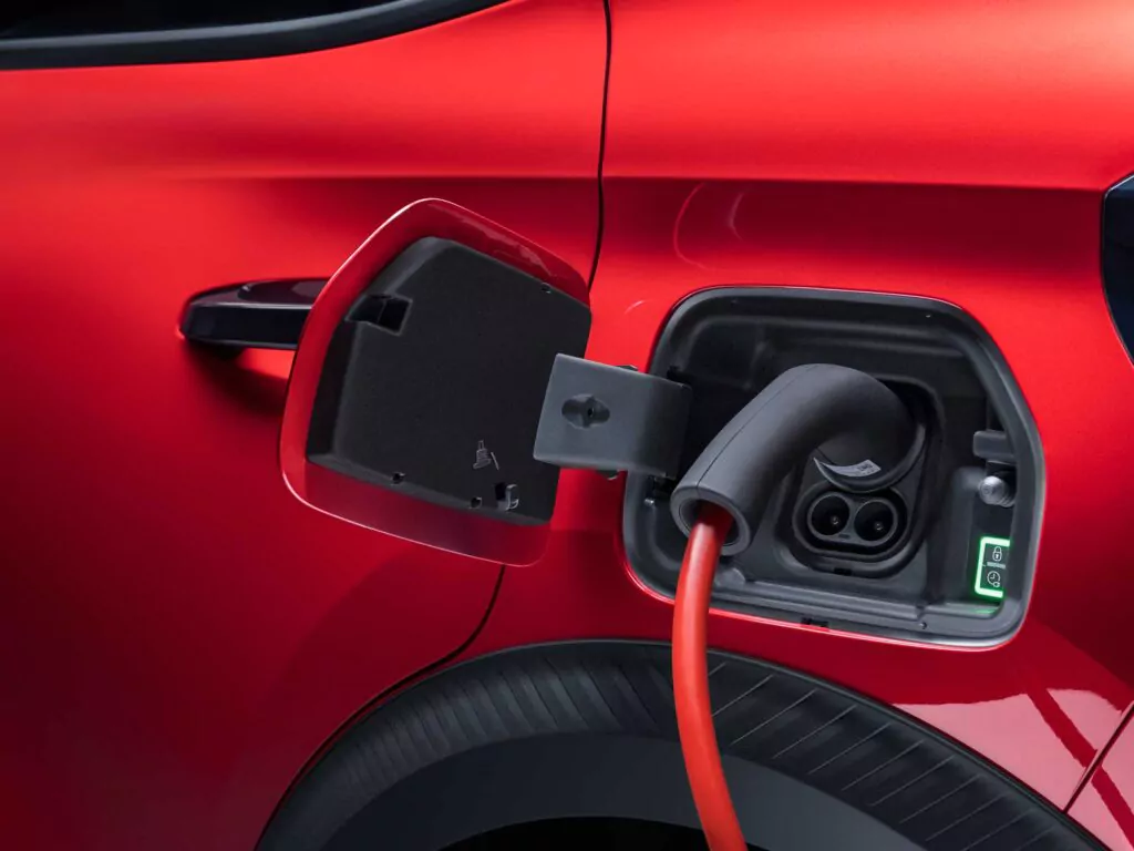 Eine Nahaufnahme eines roten Elektroautos, das aufgeladen wird, wobei der Ladeanschluss offen ist und ein orangefarbenes Ladekabel eingesteckt ist. Das glänzende Äußere des Autos und der Ladestecker sind deutlich zu sehen.