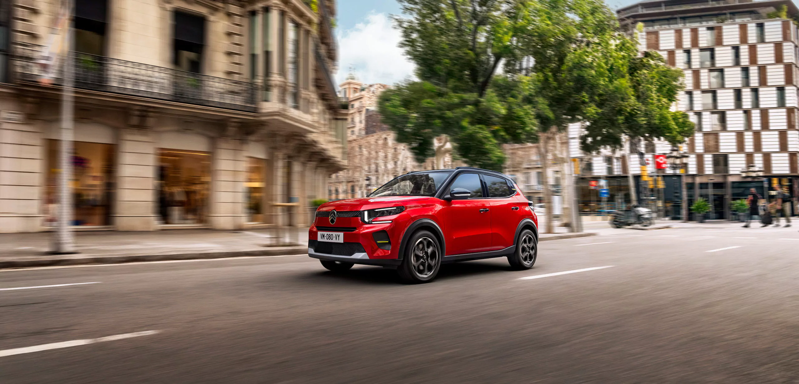 Ein roter, kompakter Geländewagen fährt eine von historischen und modernen Gebäuden gesäumte Straße entlang, wobei Bäume und verschwommene Hintergründe Bewegung suggerieren.