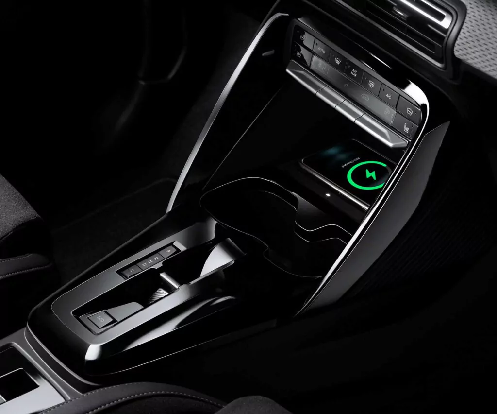 Nahaufnahme der Mittelkonsole eines modernen Autos mit Schalthebel, Becherhaltern, Bedientasten und einem Smartphone, das auf einem kabellosen Ladepad mit grünem Ladesymbol liegt.