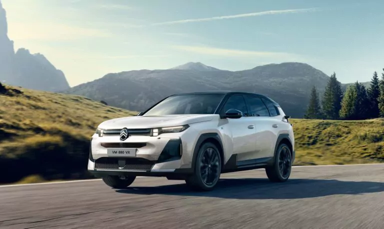 Ein weißer Citroën Geländewagen fährt auf einer asphaltierten Straße durch eine malerische Landschaft mit grasbewachsenen Hügeln, Bäumen und Bergen unter einem klaren blauen Himmel.