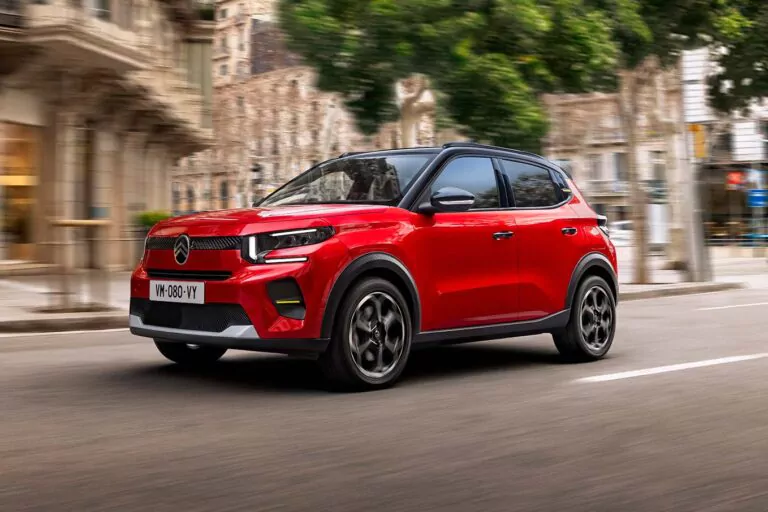 Ein leuchtend roter, kompakter Geländewagen fährt zügig eine Stadtstraße entlang, wobei Bäume und historische Gebäude im Hintergrund verschwimmen und ein Gefühl der Bewegung vermitteln. Das Fahrzeug hat ein modernes Styling und schwarze Akzente.