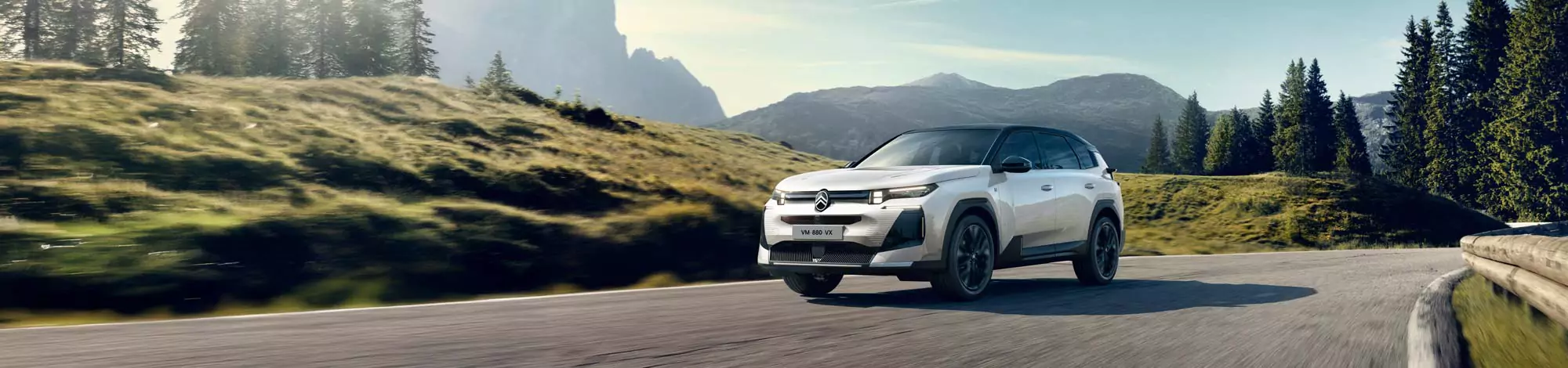 Ein weißer Geländewagen fährt auf einer kurvenreichen Bergstraße, umgeben von grünen Hügeln, Bäumen und fernen Bergen unter einem klaren blauen Himmel.