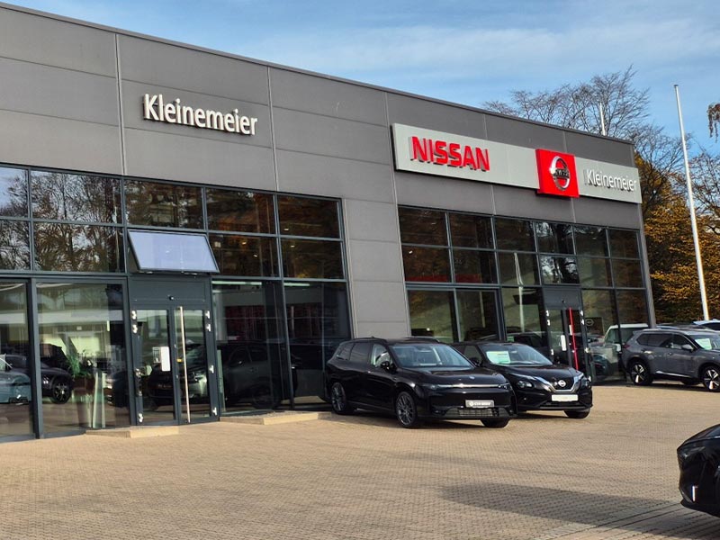 Das Gebäude eines Autohauses mit großen Fenstern, auf deren Fassade die Namen Nissan und Kleinemeier zu lesen sind. Mehrere schwarze Autos sind vor dem Autohaus geparkt. Im Hintergrund sind Bäume zu sehen.