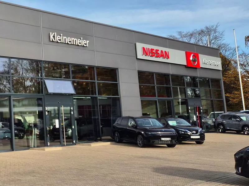 Das Gebäude eines Autohauses mit großen Fenstern, auf deren Fassade die Namen Nissan und Kleinemeier zu lesen sind. Mehrere schwarze Autos sind vor dem Autohaus geparkt. Im Hintergrund sind Bäume zu sehen.