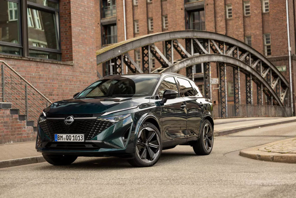 Ein dunkelgrüner Nissan Qashqai-Geländewagen ist auf einer Straße in der Nähe von Backsteingebäuden und einer Metallbrücke geparkt, mit modernen und historischen Architekturelementen im Hintergrund.