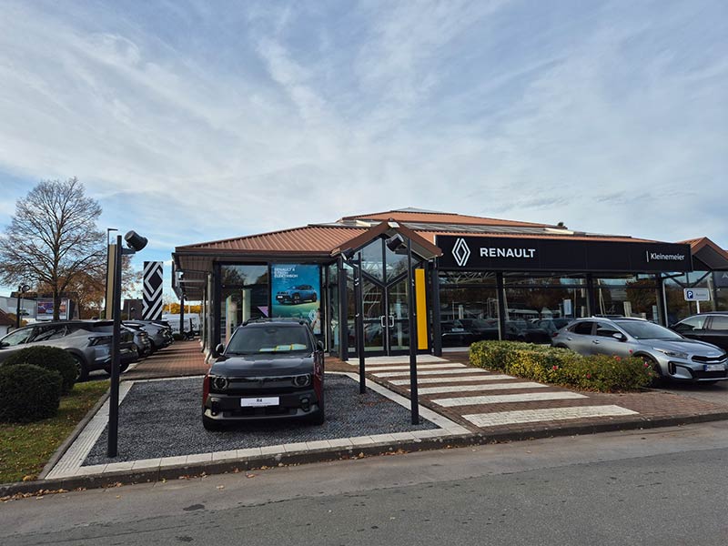 Auto Mattern: Renault Autohaus mit einem gläsernen Ausstellungsraum, mehreren Autos im Inneren und einem, das draußen neben einem Wegweiser geparkt ist. Der Himmel ist teilweise bewölkt und Bäume mit Herbstlaub umgeben das Gebäude.