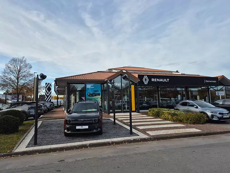 Auto Mattern: Renault Autohaus mit einem gläsernen Ausstellungsraum, mehreren Autos im Inneren und einem, das draußen neben einem Wegweiser geparkt ist. Der Himmel ist teilweise bewölkt und Bäume mit Herbstlaub umgeben das Gebäude.