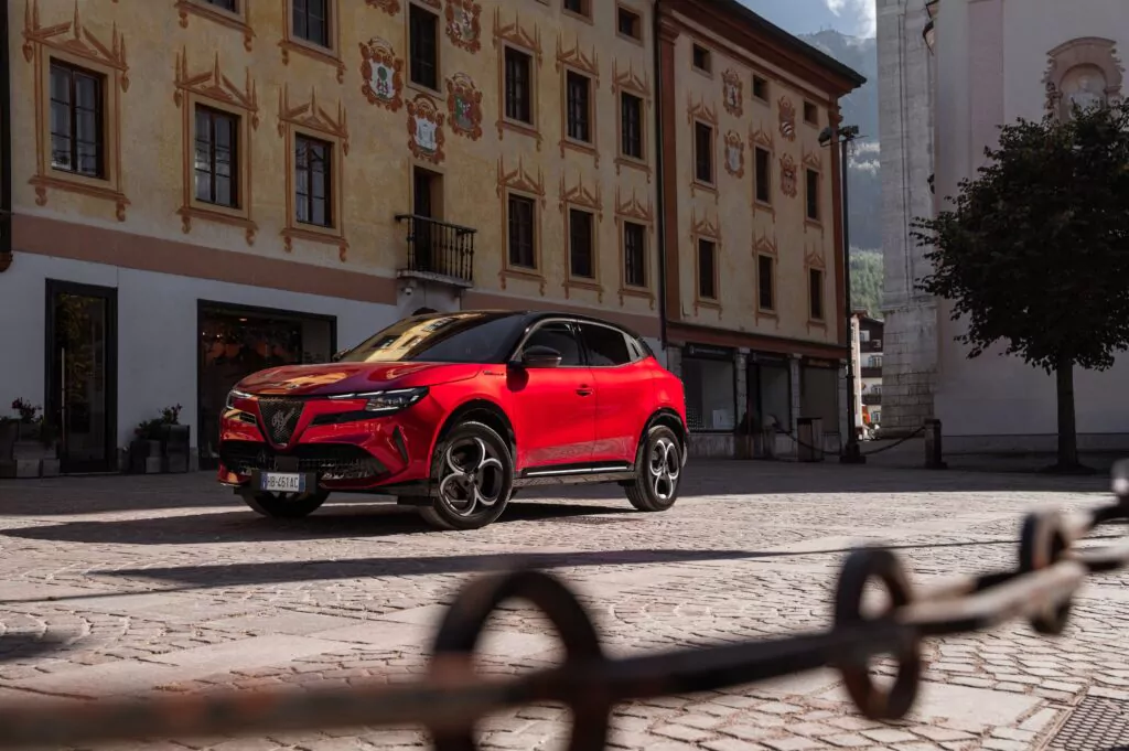 Ein roter Alfa Romeo Geländewagen parkt auf einer Kopfsteinpflasterstraße vor einem historischen Gebäude mit verschnörkelten gelben Wänden und dekorativen Fenstern. Die Szene ist hell und friedlich, und im Hintergrund sind die Berge zu sehen.