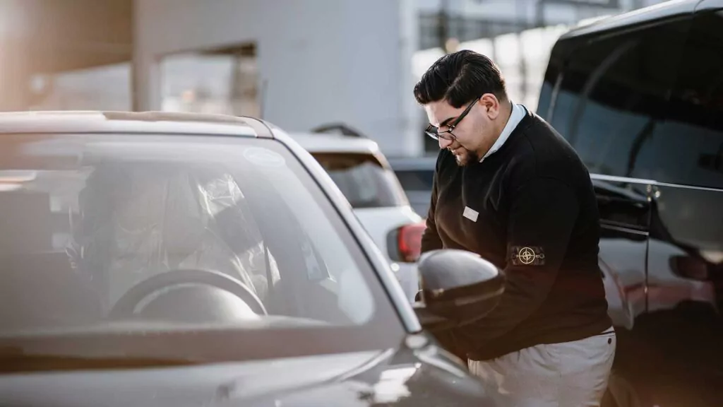 Ein Mann mit Brille und schwarzem Pullover prüft oder reinigt den Seitenspiegel eines geparkten silbernen Autos in einem Autohaus oder auf einem Parkplatz, wobei andere Fahrzeuge und ein Gebäude im Hintergrund zu sehen sind.