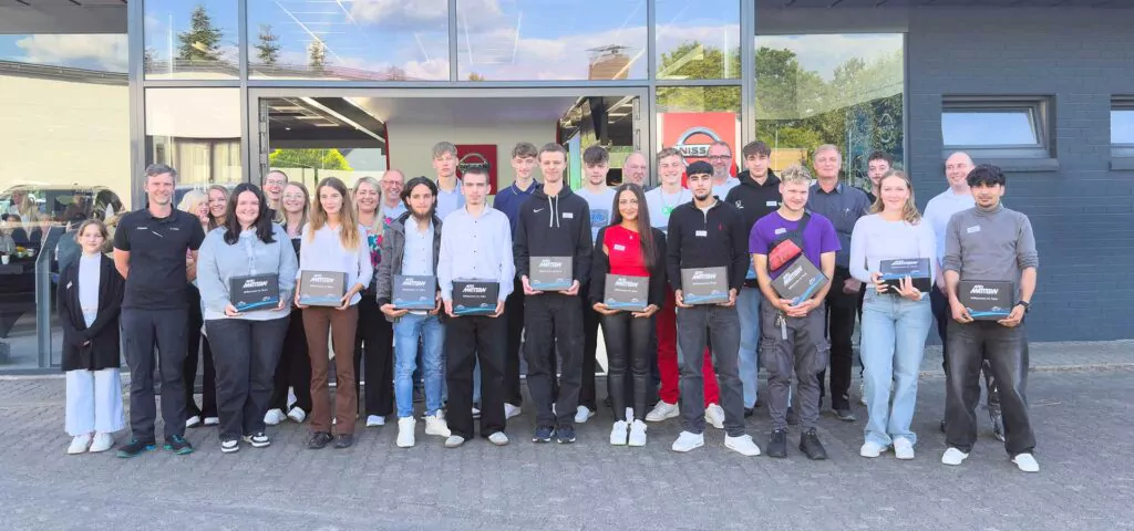 Eine Gruppe junger Erwachsener und mehrere ältere Erwachsene, möglicherweise Teilnehmer der Ausbildungswerkstatt oder künftige KFZ-Mechatroniker, stehen in zwei Reihen vor einem Gebäude, viele von ihnen halten Schachteln in der Hand und lächeln in die Kamera. In großen Fenstern spiegeln sich Bäume im Hintergrund.