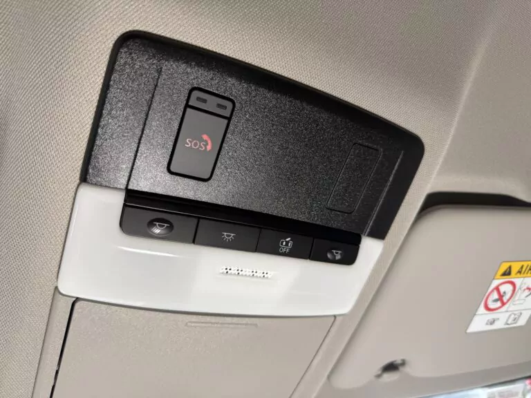 Nahaufnahme der Dachkonsole eines Autos mit einer SOS-Notfalltaste, zwei weiteren Tasten mit Symbolen und Airbag-Warnaufklebern, die teilweise auf der Sonnenblende sichtbar sind.
