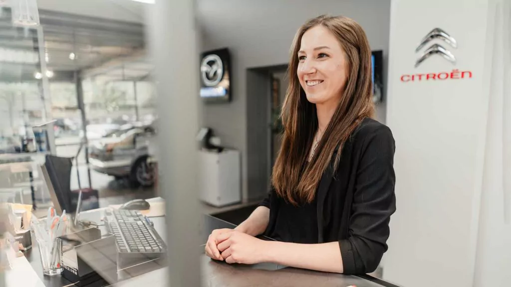 Eine Frau mit langen braunen Haaren und schwarzem Blazer steht lächelnd hinter dem Empfangstresen eines modernen Autohauses, in dem im Hintergrund die Logos von Citroën und Mazda zu sehen sind.