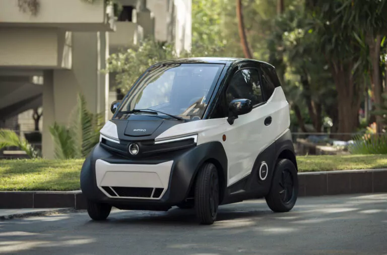 Ein kompaktes, zweitüriges Elektroauto mit schwarzem Dach und weißer Karosserie ist auf einer Straße in einer städtischen Umgebung geparkt, umgeben von Grünflächen und Gebäuden.