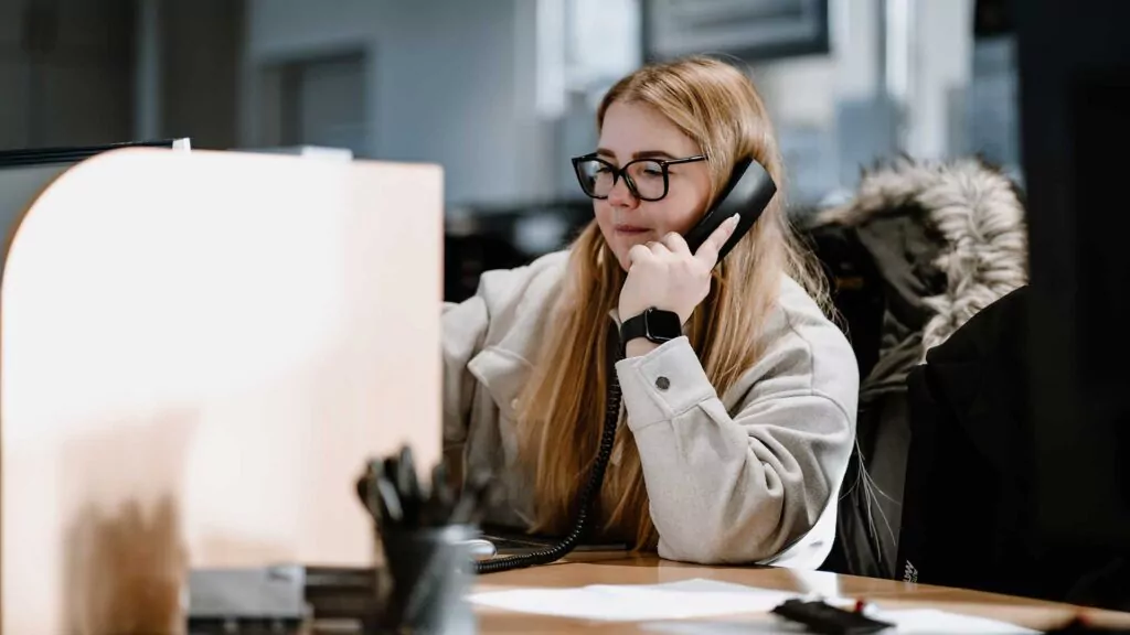 Eine Frau mit langen blonden Haaren und Brille sitzt an einem Schreibtisch und spricht in ein Festnetztelefon. Sie trägt einen hellen Mantel und arbeitet in einer Büroumgebung, in der Papiere und ein Stifthalter auf dem Schreibtisch zu sehen sind.