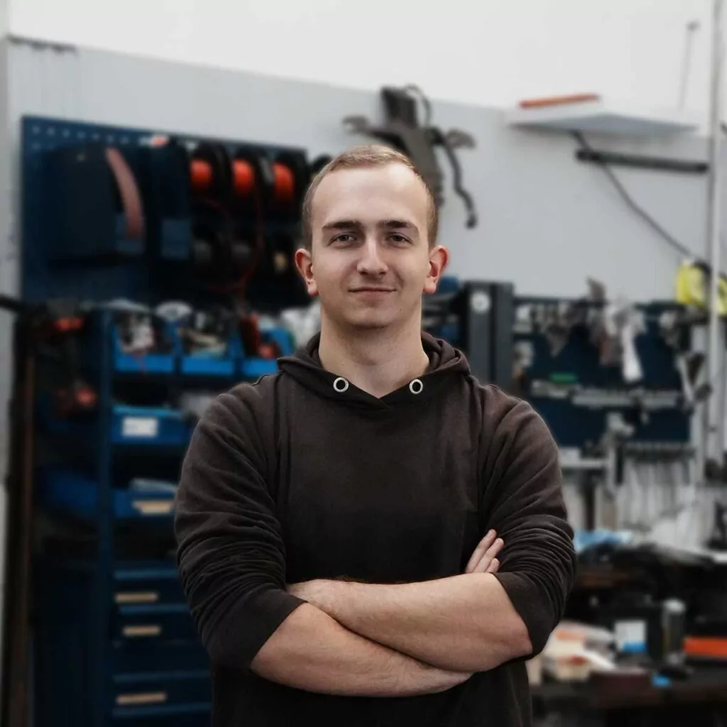 Ein junger Mann mit kurzen Haaren steht selbstbewusst mit verschränkten Armen in einer Werkstatt, umgeben von Werkzeugen, Regalen und Geräten.