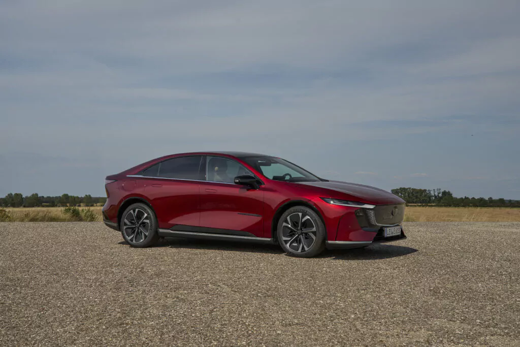 Ein schlankes, rotes, modernes Elektroauto, geparkt auf einer Kiesfläche mit einem offenen Feld und blauem Himmel im Hintergrund. Das Auto hat ein stromlinienförmiges Design und schwarze Zierelemente.