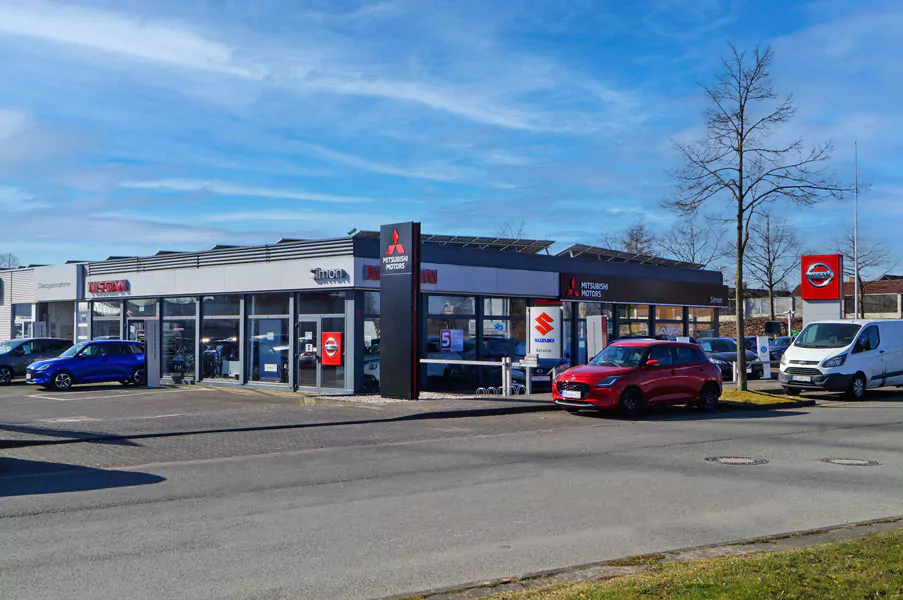 DSC06165_bearbeitet_3_klein Ein Autohaus mit Nissan-, Suzuki- und Mitsubishi-Schildern am Gebäude, mehrere Autos, die draußen geparkt sind, und ein klarer blauer Himmel über uns.