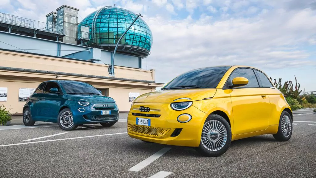 Zwei Fiat 500 Elektroautos, ein gelbes und ein blaugrünes, sind im Freien auf einer gepflasterten Straße geparkt, mit einem modernen Gebäude mit Glaskuppel und einem bewölkten Himmel im Hintergrund.