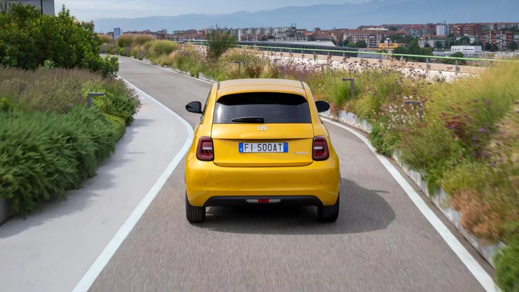 Ein gelber Fiat 500 fährt auf einer begrünten Dachterrassenstraße, umgeben von grünen Pflanzen und Blumen, während im Hintergrund eine Stadtlandschaft und Berge zu sehen sind.