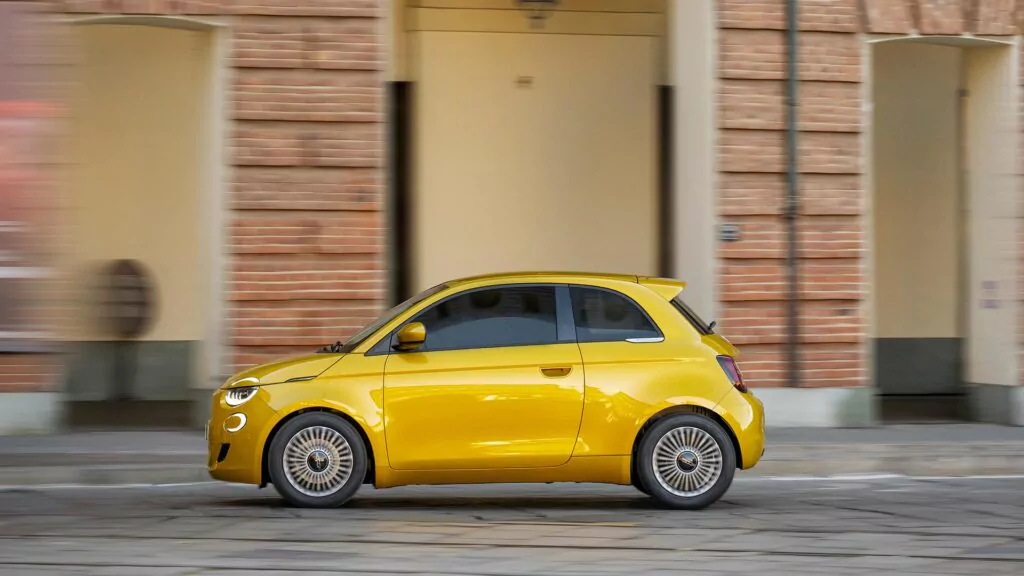 Ein leuchtend gelber Fiat 500 fährt auf einer Stadtstraße neben einem Gebäude mit Backsteinmauern und gewölbten Türöffnungen.