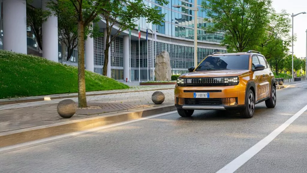 Ein bronzefarbener Fiat Grande Panda fährt an einem sonnigen Tag eine von Bäumen und modernen Glasgebäuden gesäumte Stadtstraße entlang. Das Fahrzeug bewegt sich auf der rechten Spur, während in der Nähe ein steinerner Bürgersteig und Straßenlampen zu sehen sind.