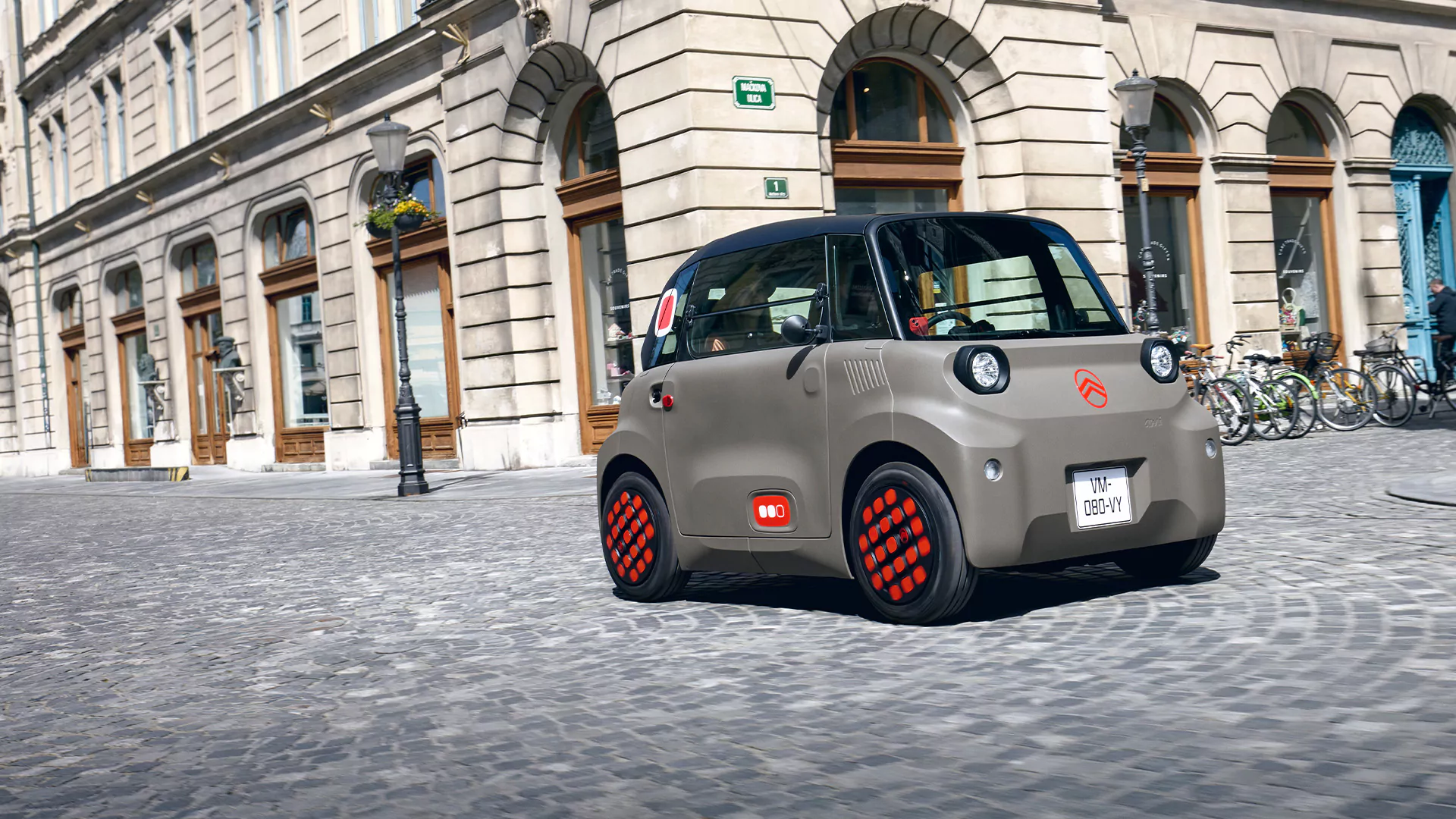 Ein kleines, graues Elektroauto vom Typ Citroën Ami mit roten Radakzenten – bekannt für seine beeindruckende Reichweite – fährt auf einer Kopfsteinpflasterstraße in einer historischen europäischen Stadt, die von Steinhäusern und geparkten Fahrrädern gesäumt ist.