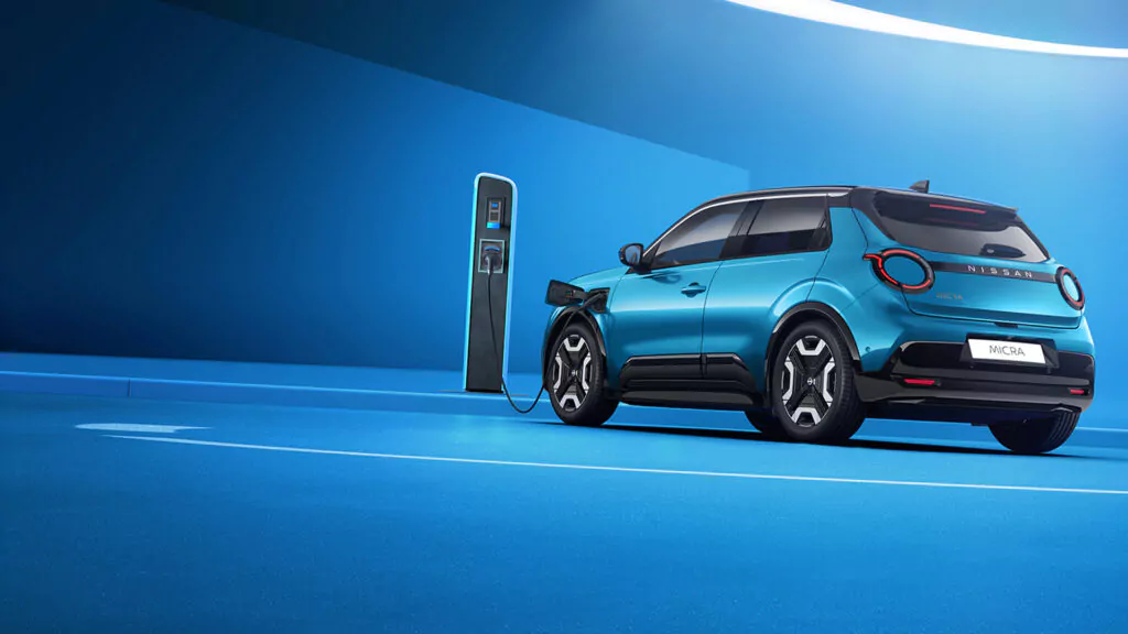 Ein blauer Nissan MICRA Engage mit Elektro-Förderung ist in einer modernen, blau getönten Ladestation geparkt und an eine Ladesäule für Elektrofahrzeuge angeschlossen. Die Szene ist elegant und futuristisch.