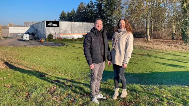 Zwei Personen stehen lächelnd auf einer Wiese vor einem modernen Gebäude mit der Aufschrift Add Matter. Im Hintergrund sind Bäume und herabgefallene Blätter unter einem klaren blauen Himmel zu sehen.