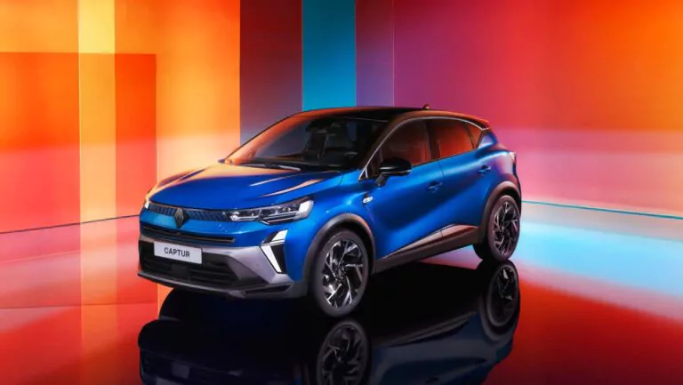 Ein blauer Renault Captur Geländewagen, der in einem Innenraum vor einem farbenfrohen, geometrischen Hintergrund mit roten, orangen und blauen Farbtönen geparkt ist. Das Auto ist angewinkelt, um seine Front und Seite zu zeigen.