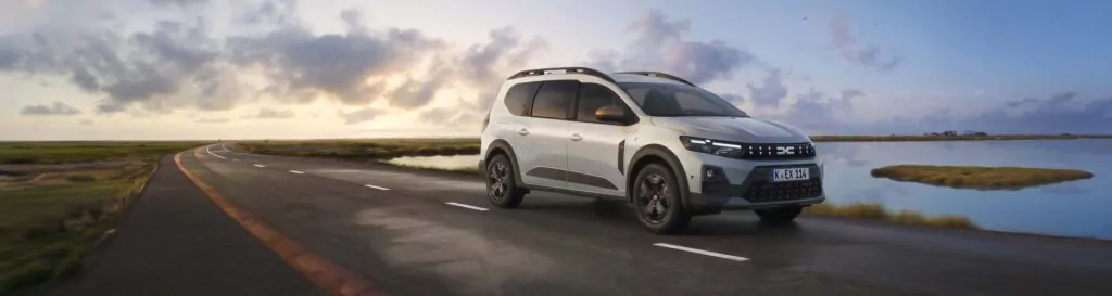 Ein silberner Geländewagen, möglicherweise ein Dacia Jogger Essential Leasing, fährt bei Sonnenaufgang oder Sonnenuntergang auf einer asphaltierten Straße an einem ruhigen See entlang, während sich der Himmel mit dramatischen Wolken füllt und sich Graslandschaften in der Ferne erstrecken.