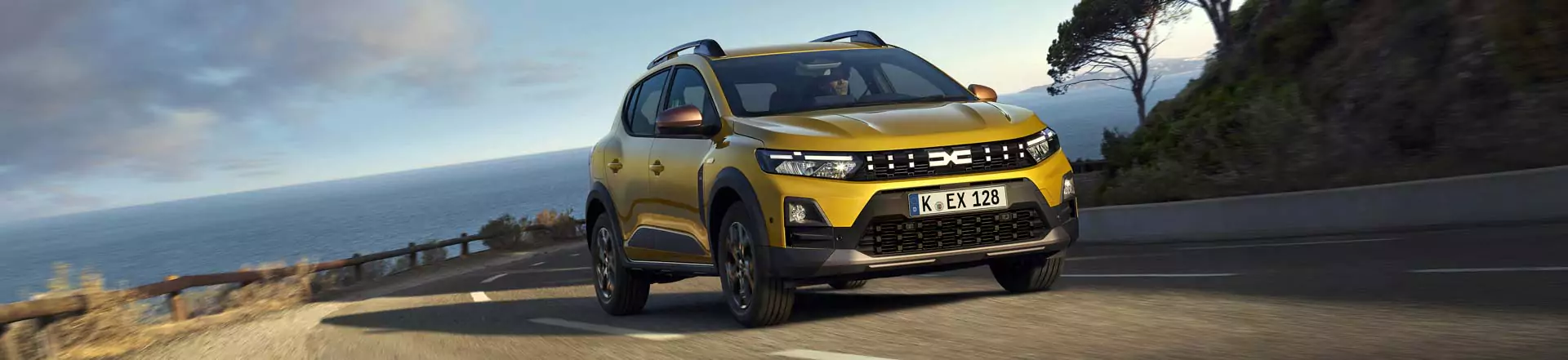 Ein gelber Dacia Sandero Essential fährt auf einer kurvenreichen Küstenstraße mit dem Meer und den Klippen im Hintergrund unter einem teilweise bewölkten Himmel.