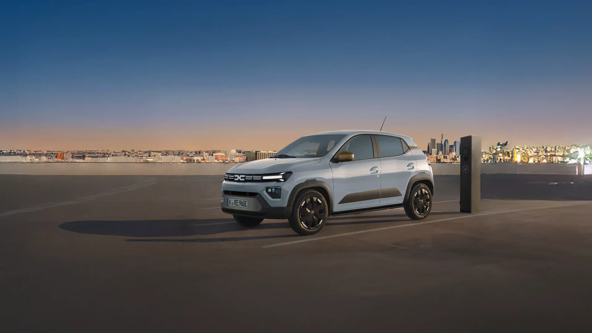 Ein kleines silbernes Elektroauto parkt und lädt an einer Ladestation auf einem überdachten Parkplatz bei Sonnenuntergang, während im Hintergrund die Skyline der Stadt zu sehen ist.
