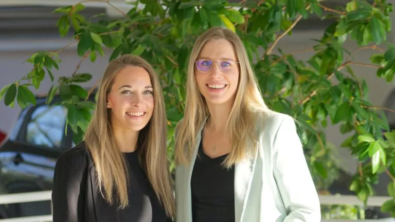 Zwei Frauen mit langen blonden Haaren stehen lächelnd vor grünen Blattpflanzen. Die eine trägt ein schwarzes Outfit, die andere einen hellen Blazer und eine Brille. Das Sonnenlicht erhellt ihre Gesichter. Im Hintergrund sind ein Auto und das Innere eines Gebäudes zu sehen.