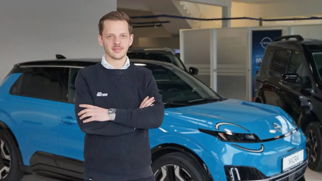 Ein Mann mit kurzen Haaren steht mit verschränkten Armen vor einem hellblauen Auto in einem Ausstellungsraum. Er trägt einen schwarzen Pullover und eine Uhr und wirkt selbstbewusst. Im Hintergrund sind andere Fahrzeuge und Ausstellungsräume zu sehen.