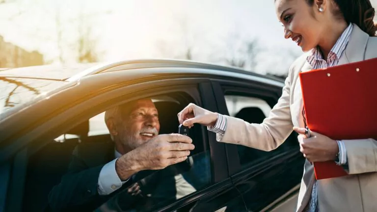 Eine lächelnde Frau übergibt einem Mann, der auf dem Fahrersitz eines Autos sitzt, die Autoschlüssel; sie hält eine rote Mappe in der Hand, und beide wirken glücklich, was auf einen erfolgreichen Autoverkauf oder eine erfolgreiche Vermietung hindeutet.