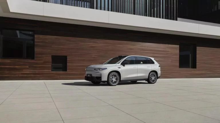 Ein silberner Leapmotor C10 ist auf einer großen, leeren Betonfläche vor einem modernen Gebäude mit einer holzgetäfelten Wand und Glasfenstern geparkt. Die Fläche ist gut beleuchtet, und auf dem Boden liegen deutliche Schatten.