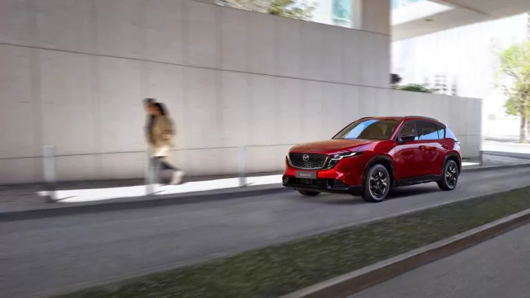 Ein roter Geländewagen fährt auf einer Stadtstraße neben einem Bürgersteig, auf dem eine Person in einem hellbraunen Mantel an einem modernen Gebäude vorbeigeht.